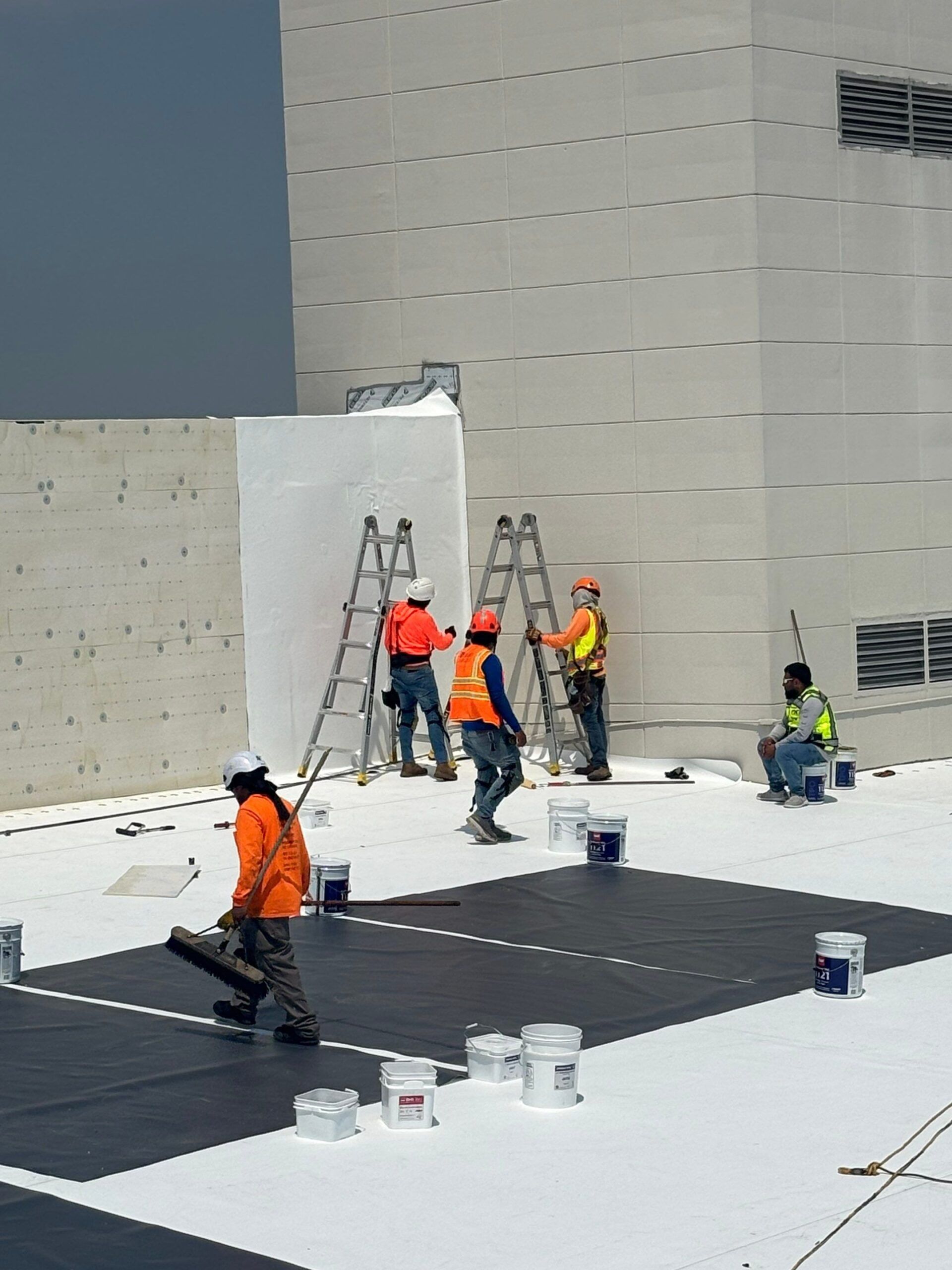Construction workers installing roofing on a building. They are wearing safety vests and using ladders.