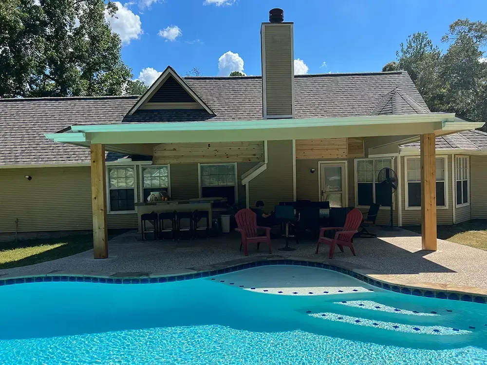 Poolside patio with a light-colored cover, adjacent to a house. A pool with blue water is in the foreground.