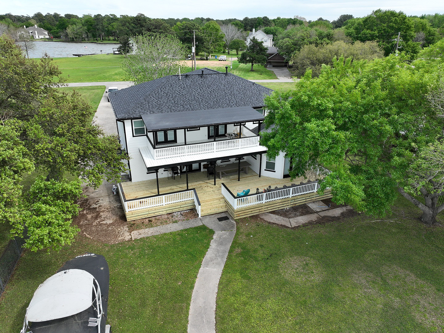 White house with black trim and a large deck, surrounded by trees and green grass.