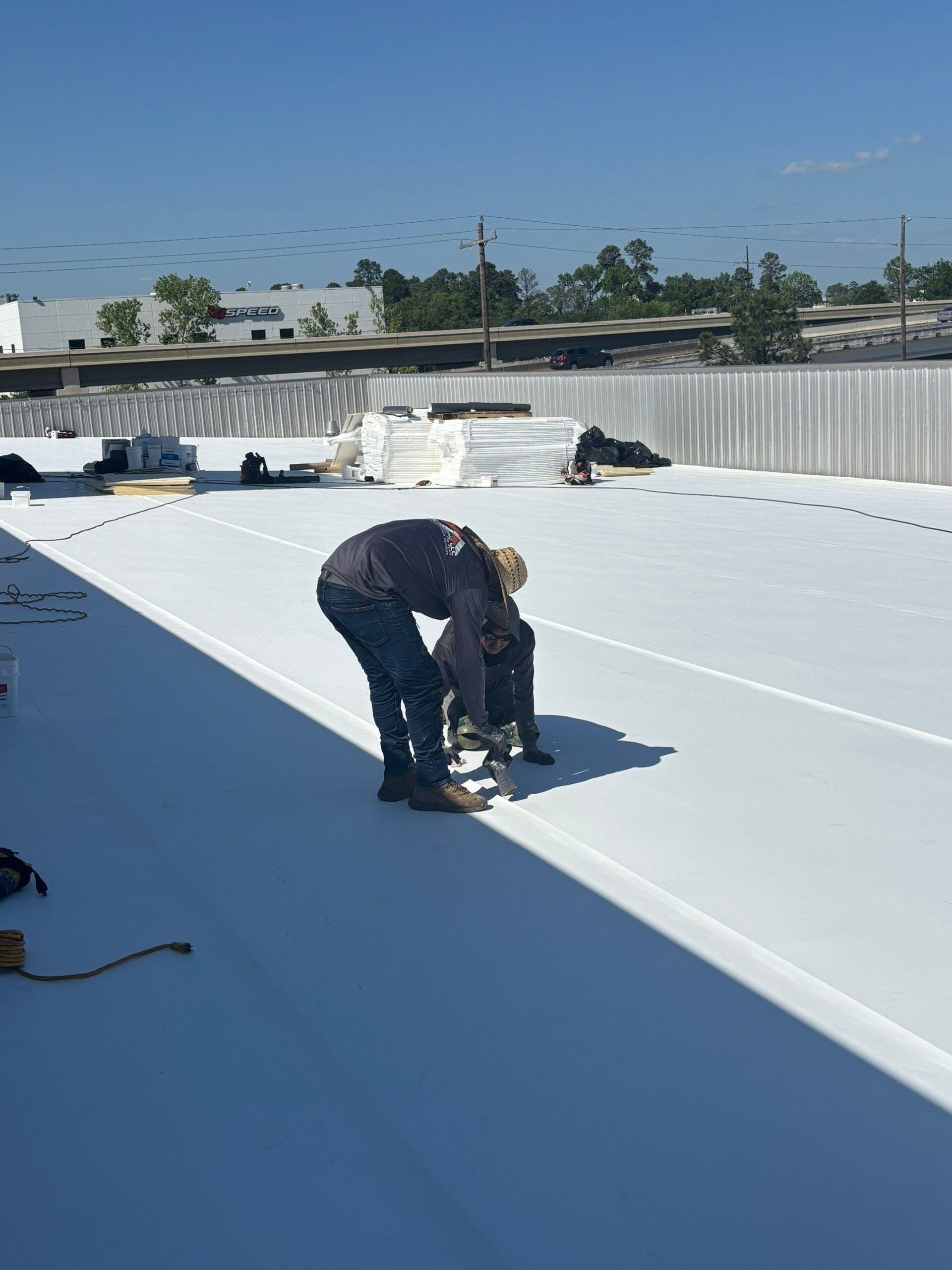 Two workers on a white rooftop; one is bending over a machine, the other stands nearby. Sunny day.