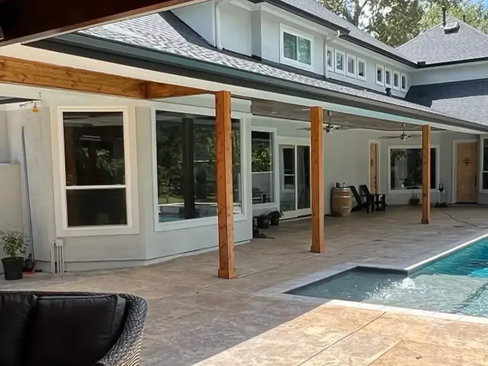 Backyard patio with pool, covered by wooden beams, with a house in the background.
