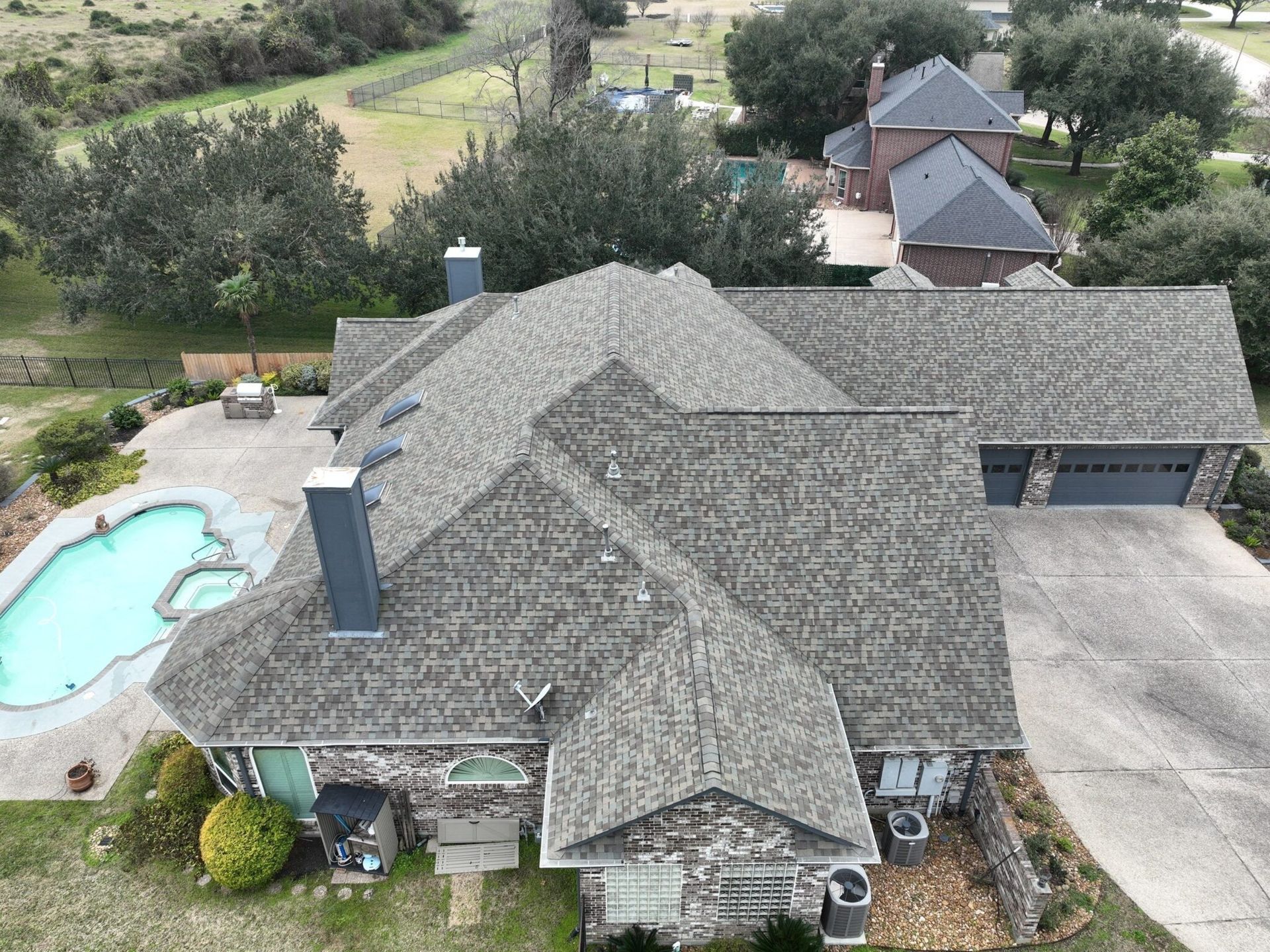 Overhead view of a house with gray roof, brick exterior, pool, and attached garage. Trees and fields surround it.