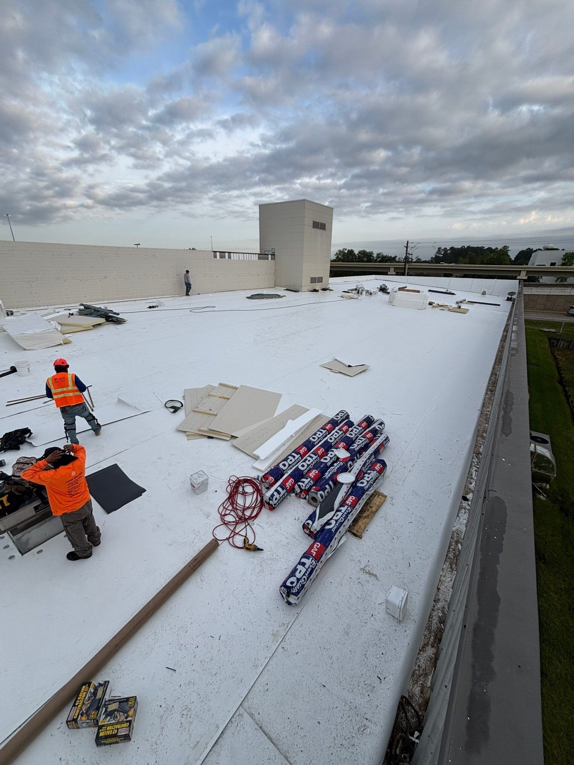 Workers installing a white roof on a building. Rolls of roofing material and tools are visible. Overcast sky.