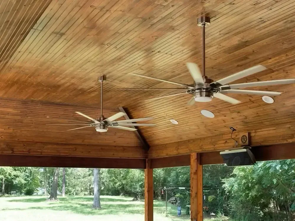 Two ceiling fans with lights in a wooden-paneled outdoor space, overlooking a green yard.
