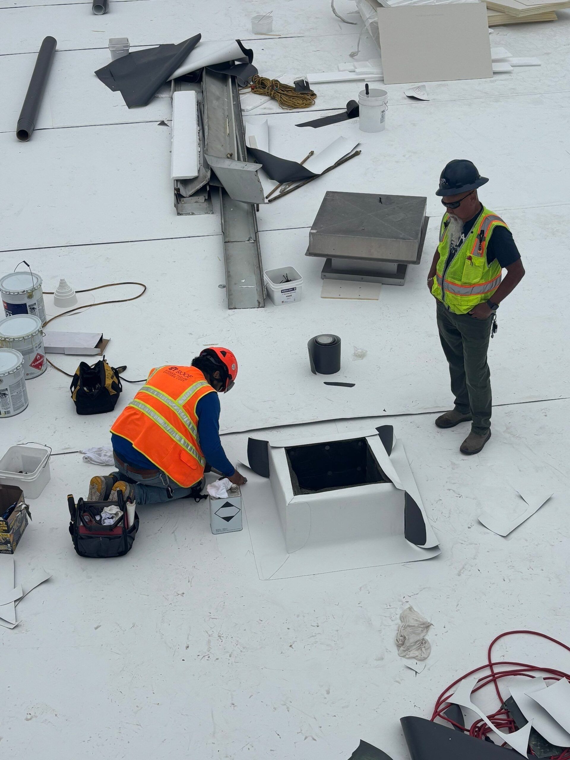 Two roofers working on a white flat roof, one kneeling near a white square structure, the other standing nearby.
