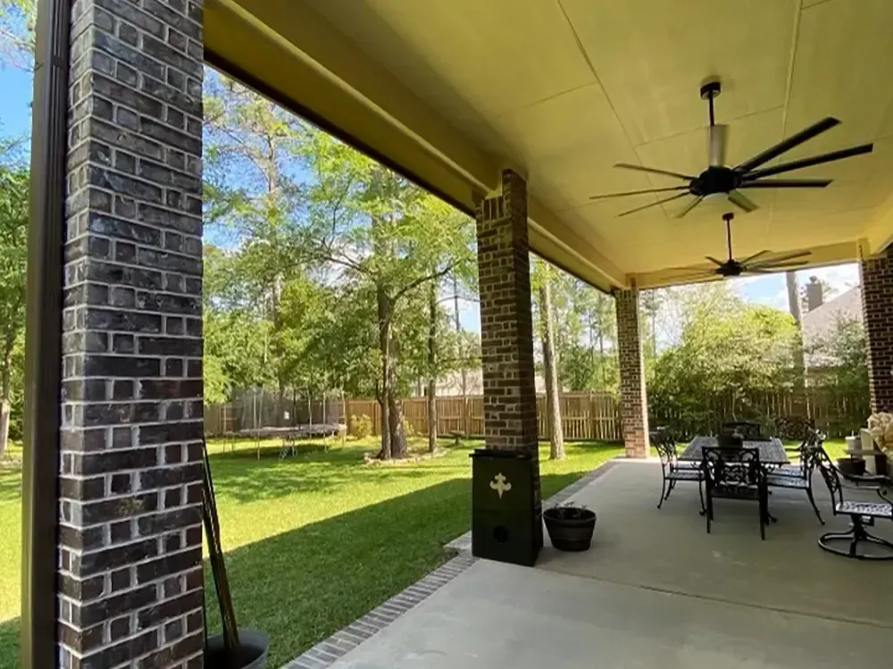 Covered patio with brick columns, ceiling fans, and a dining set overlooking a grassy yard.