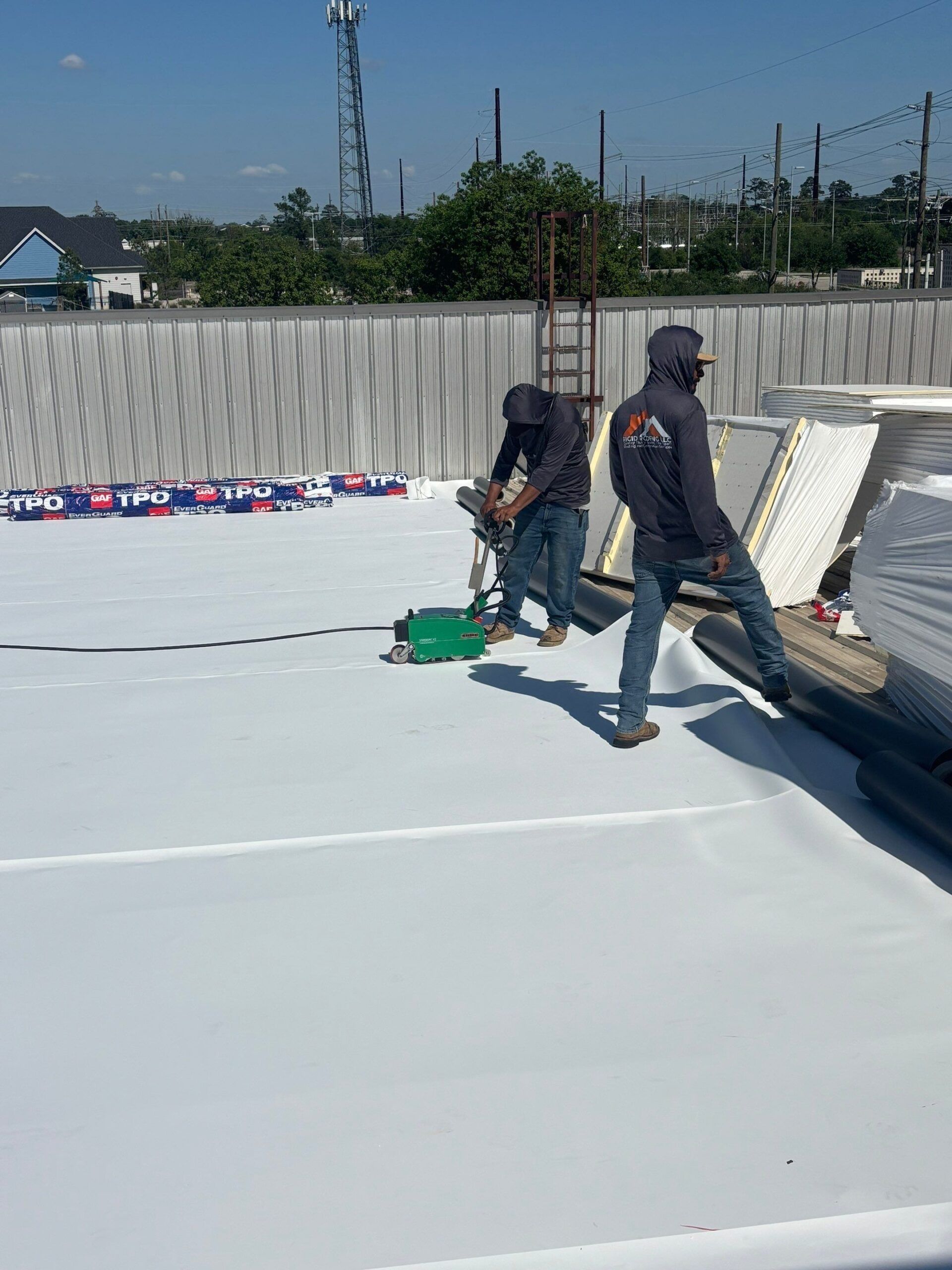 Two workers installing roofing on a sunny day. One uses a green tool; the other assists.