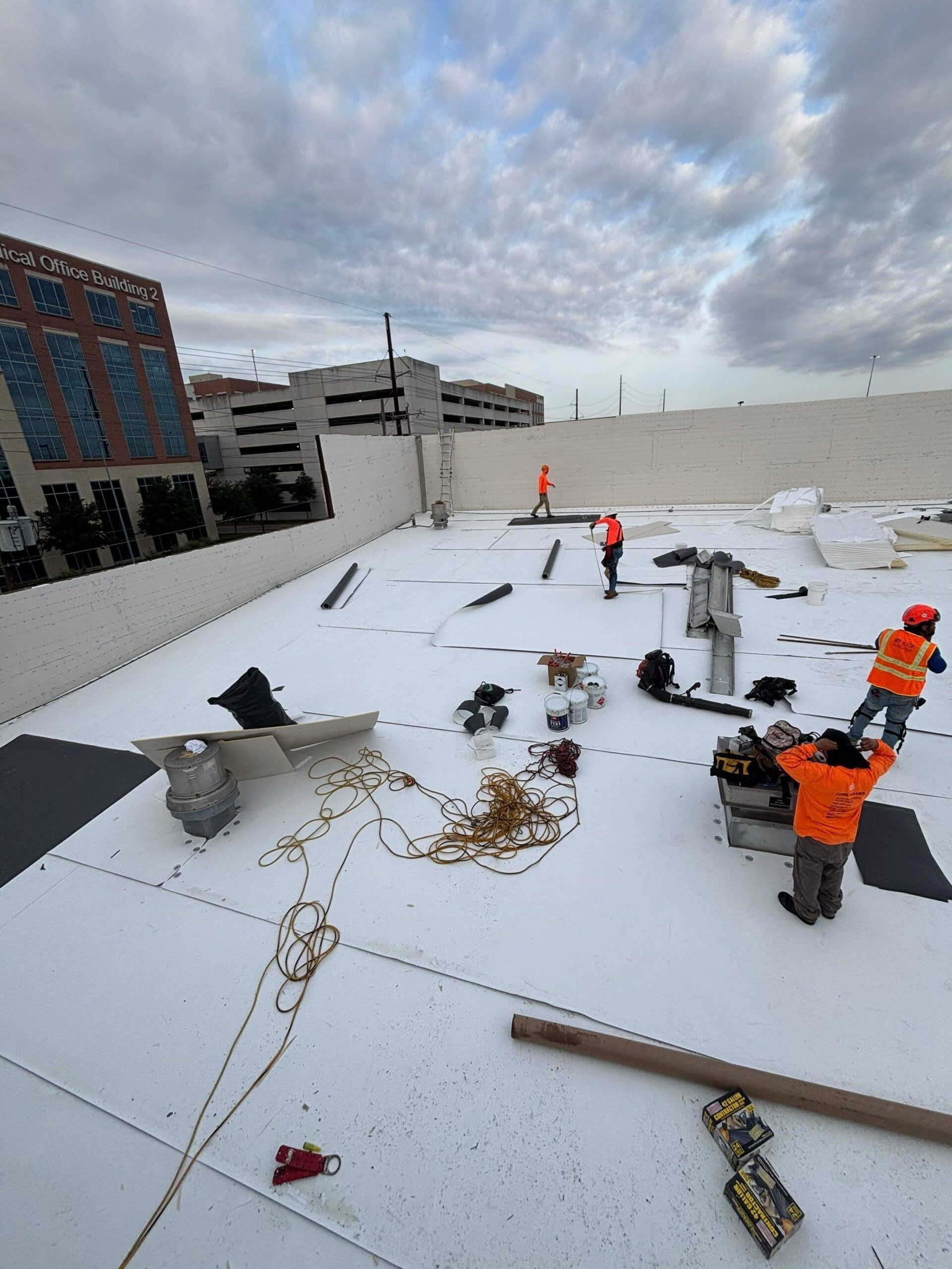 Construction workers in orange vests on a white roof, overcast sky, tools and materials scattered.
