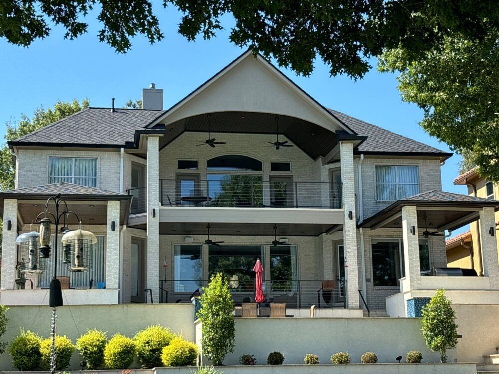 Two-story white brick home with balconies and dark roof overlooking a green yard and water on a sunny day.
