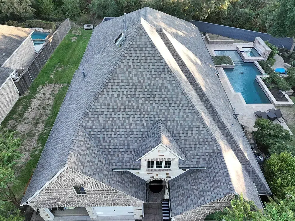 Aerial view of a large house with a grey shingled roof, a pool, and surrounding green lawn.