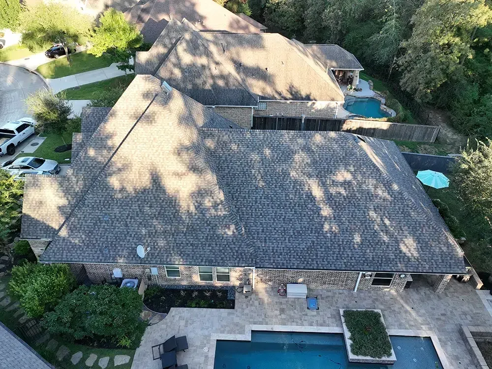 Aerial view of a large home with a pool in the backyard. Gray roof, trees, and cars in the driveway.