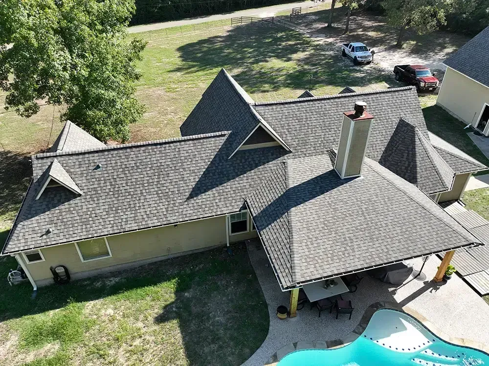 Aerial view of a house with a gray shingle roof, chimney, and a blue swimming pool in the backyard.