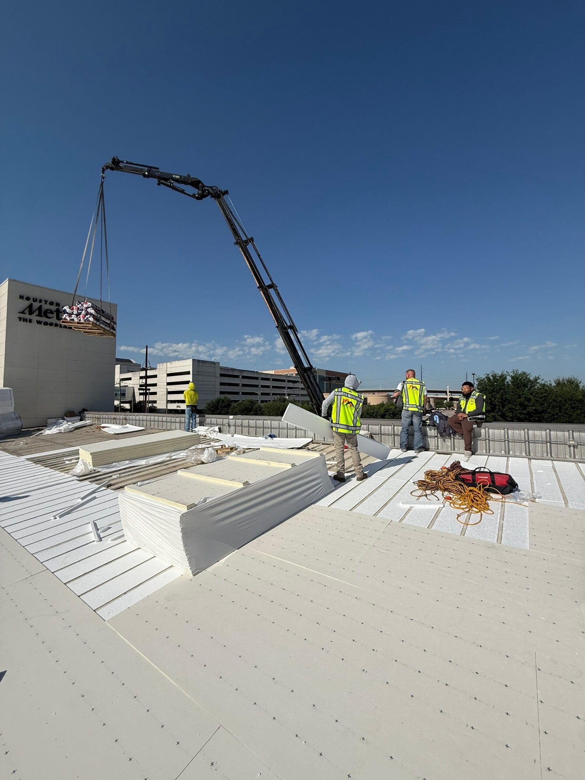 Construction workers on a roof use a crane to lift materials, with a blue sky background.