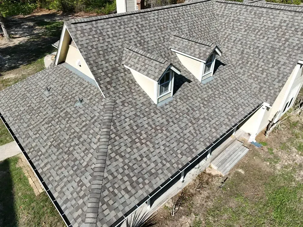 Overhead view of a house with a gray shingle roof, three dormers, and light-colored siding, surrounded by trees.