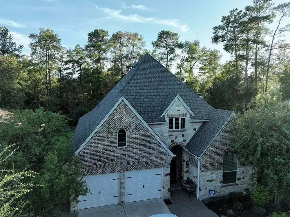 Two-story house with a gray roof and brick and stone facade, surrounded by trees.