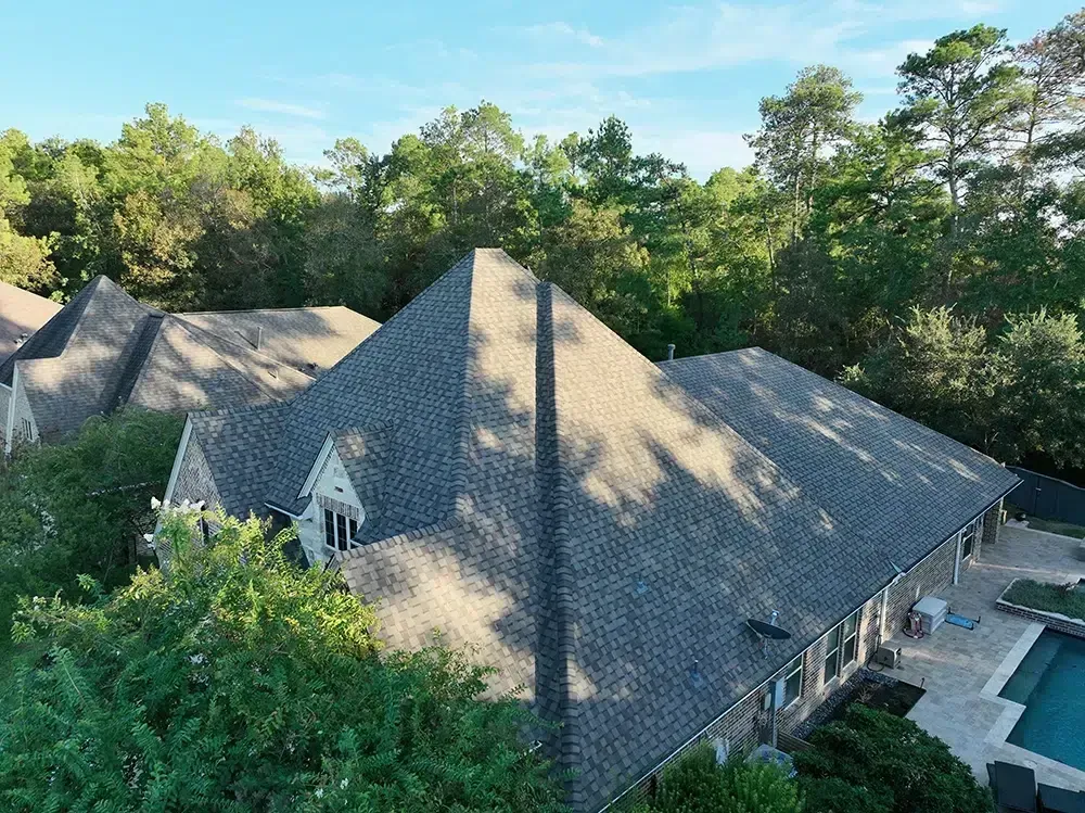 An aerial view of a house with a gray shingled roof, trees, and a pool.