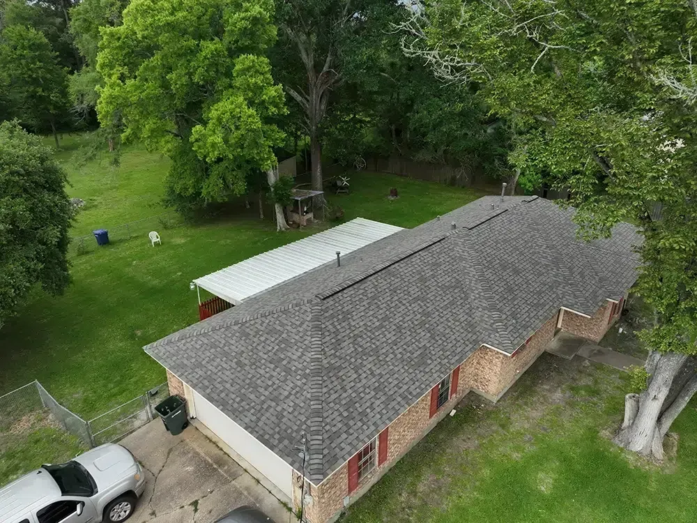 Aerial view of a single-story house with a gray roof and attached carport, surrounded by trees and green grass.