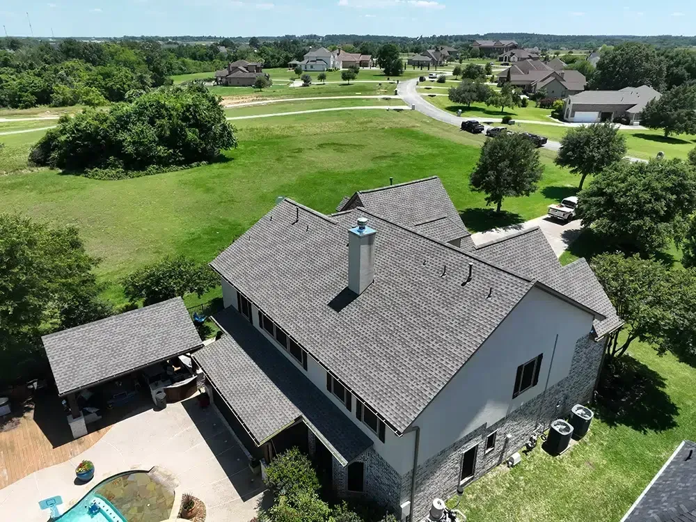 Aerial view of a gray-roofed house with a swimming pool, nestled in a green landscape with other houses and trees.
