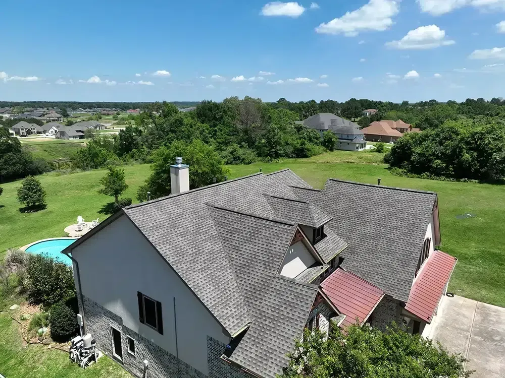 Aerial view of a house with a gray roof and a pool, surrounded by green grass and trees under a blue sky.