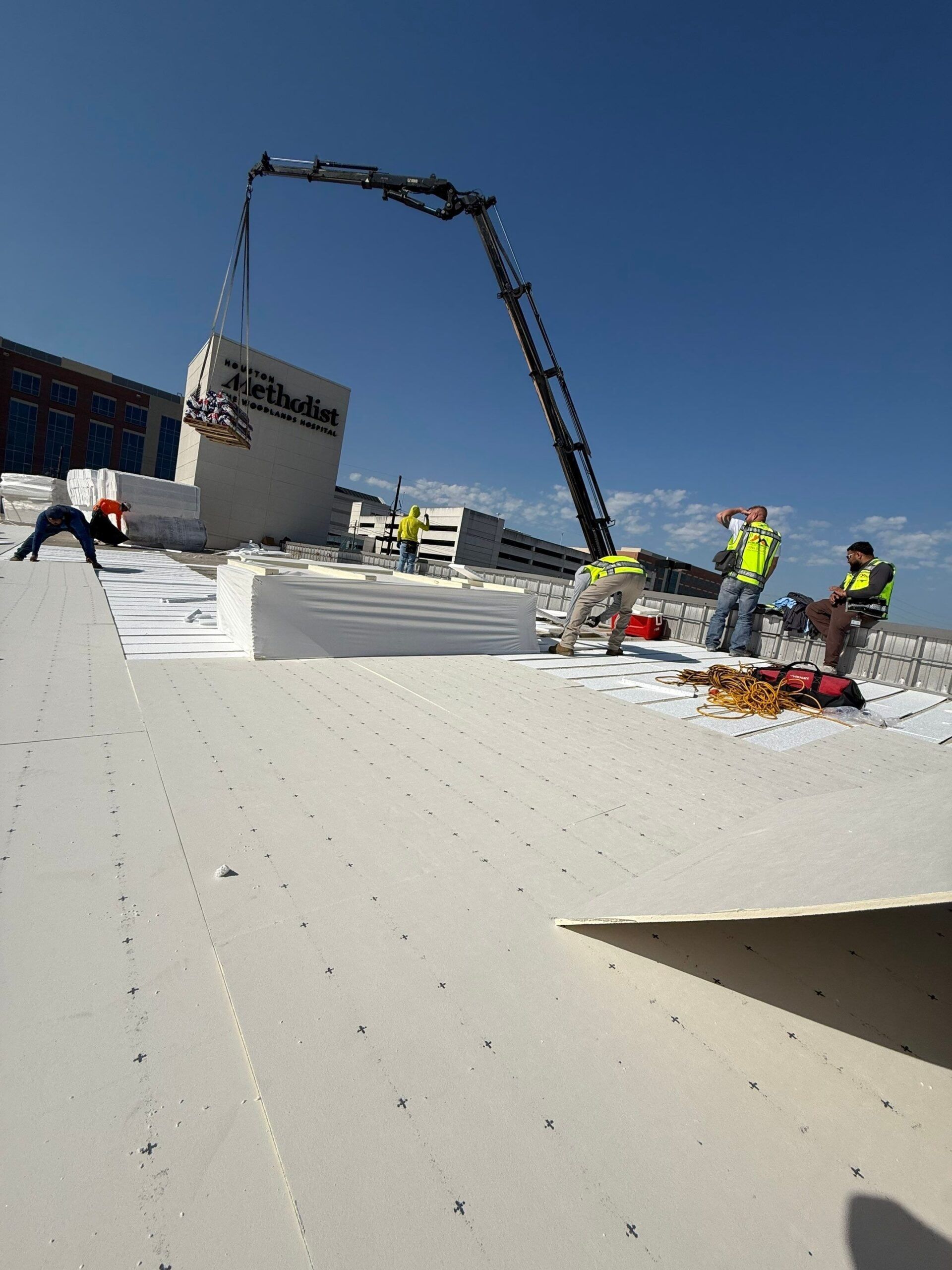 Crane lifting a large concrete block onto a rooftop; workers in safety vests; blue sky.