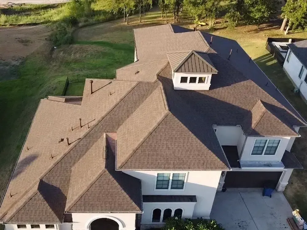 Brown shingled roof of a two-story house with dormers and attached garage, viewed from above.