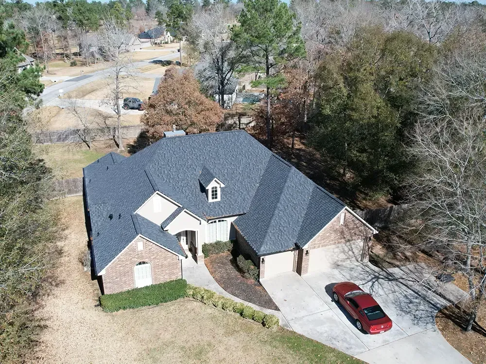 Aerial view of a house with a dark roof and a red car in the driveway, surrounded by trees.