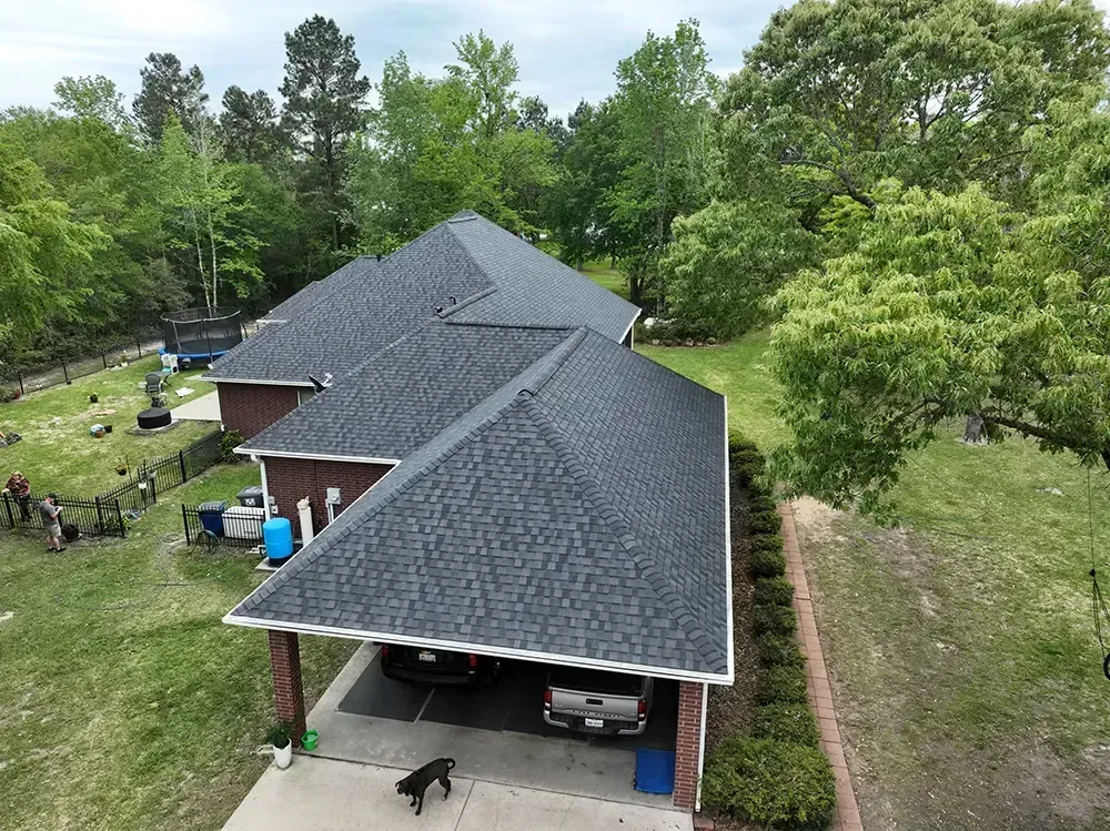 House with dark gray roof and carport, surrounded by trees and lawn. A dog stands on a driveway.