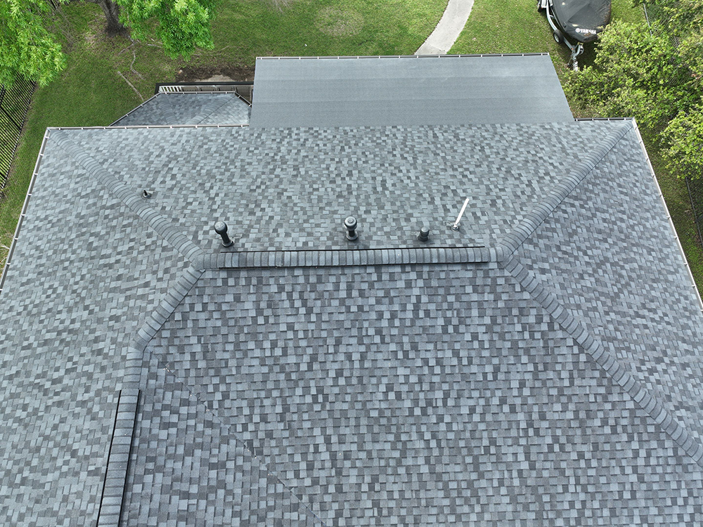Overhead view of a house roof with dark gray shingles, multiple vents, and a small section of a lighter roof.
