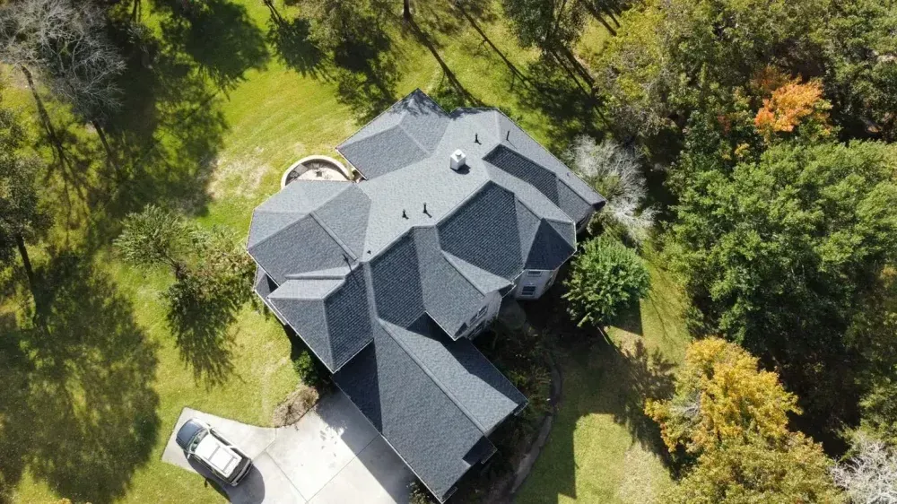 Aerial view of a dark-roofed house with multiple sections surrounded by trees and a grassy lawn.