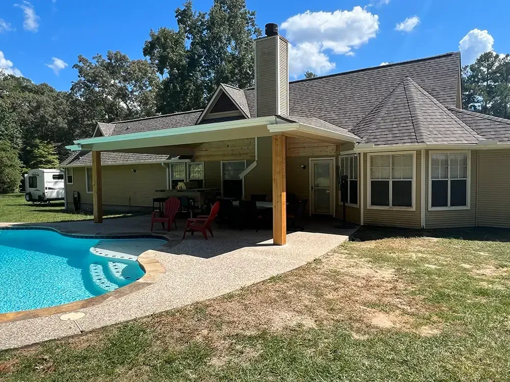 A pool with a patio and house. The patio has a cover. Trees and a camper are in the background.