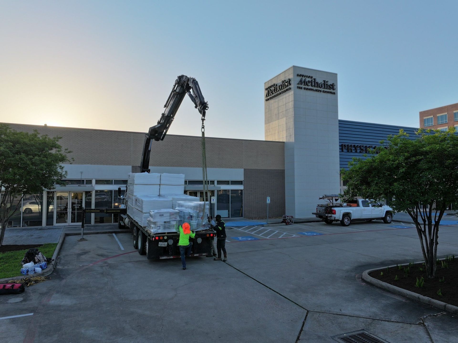 A crane lifts materials from a truck in front of a building. Two workers in vests and a pickup truck are visible.