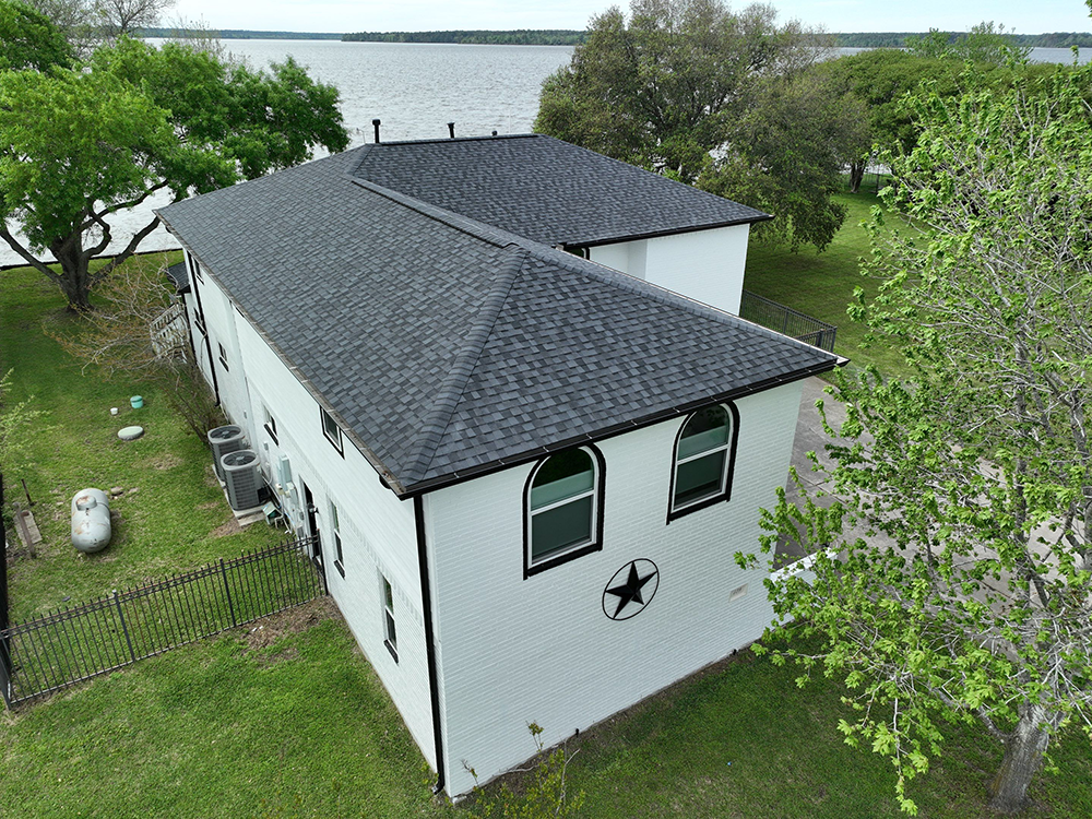 White two-story building with a black shingle roof, facing a body of water. Green grass and trees surround it.