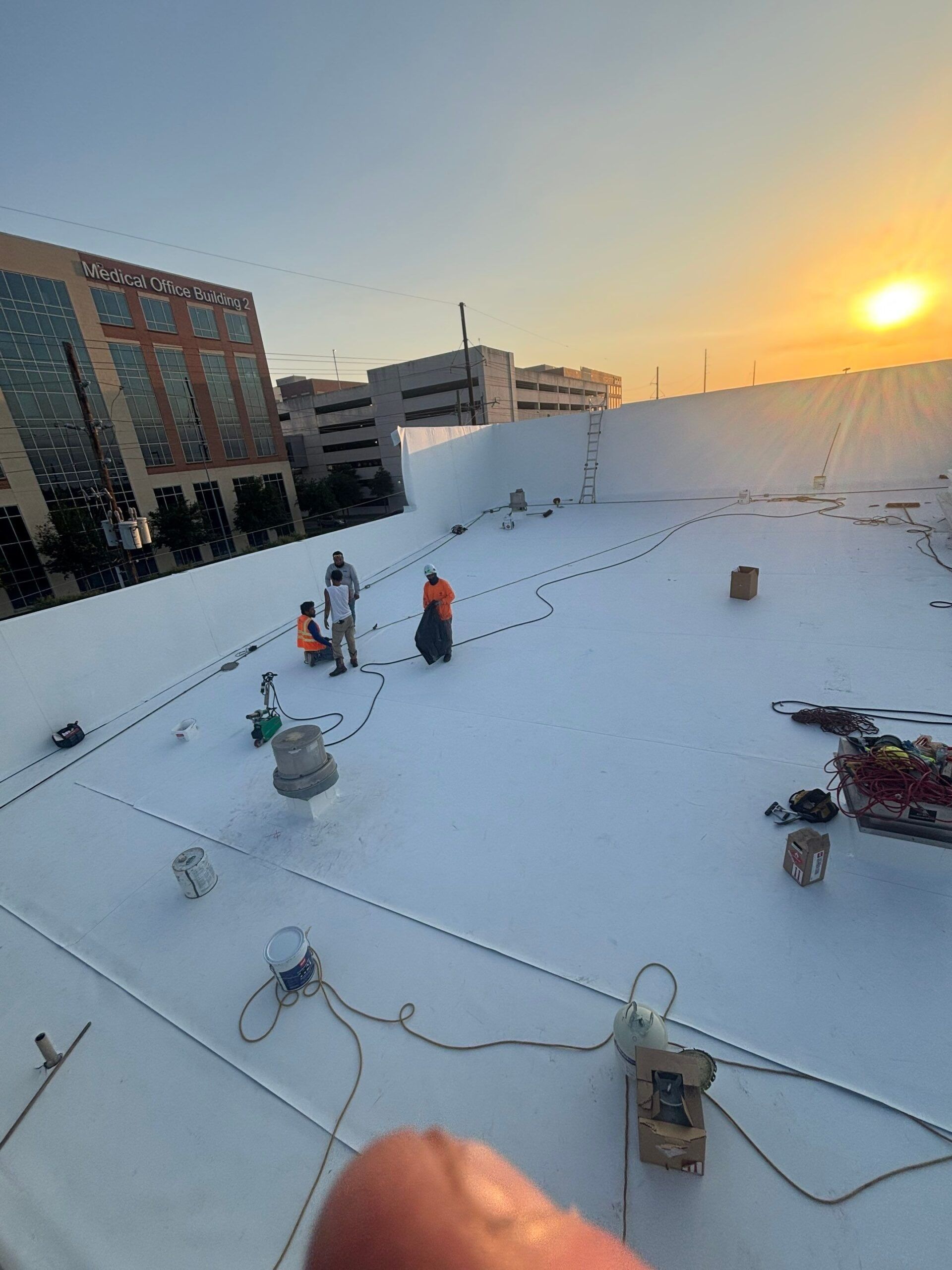 Workers on a white roof at sunset; building and cityscape in background.