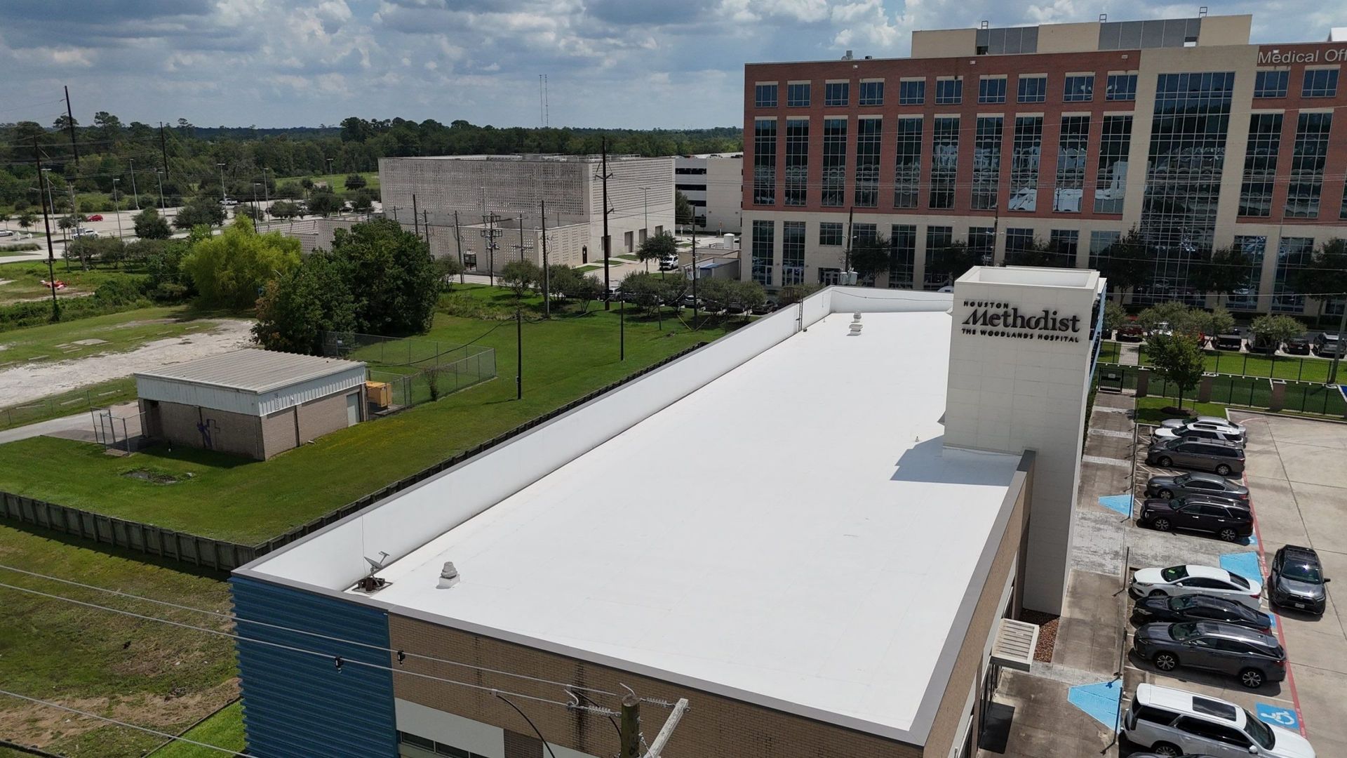Aerial view of a building with a white roof, next to a parking lot and other structures.