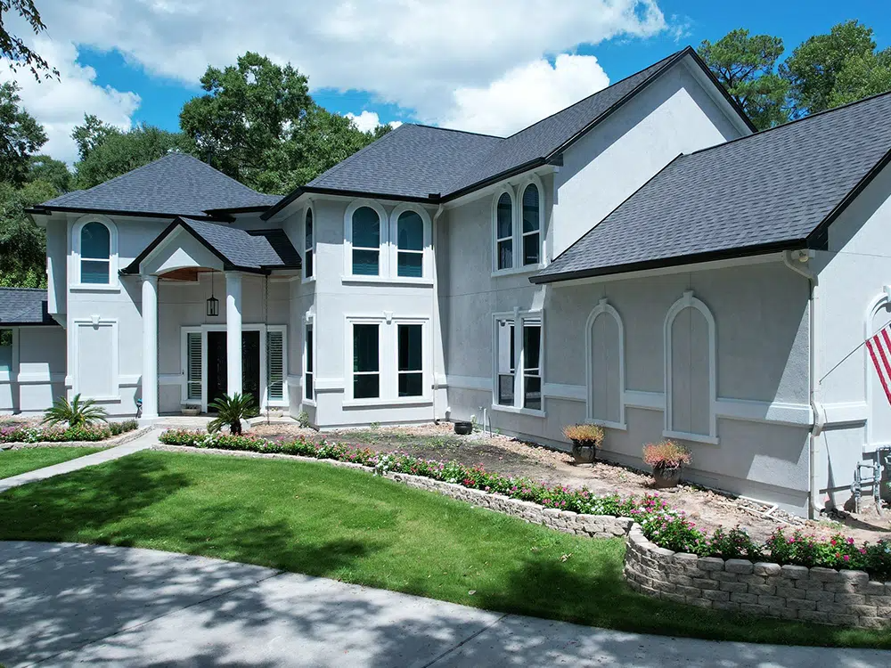 Large, light-colored house with dark roof and trim, green lawn, and landscaping.