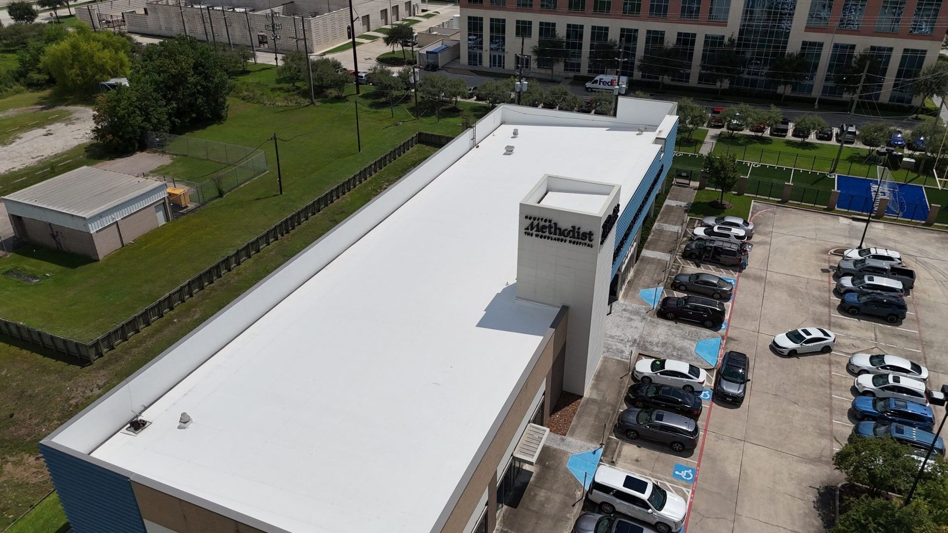 Aerial view of a white-roofed commercial building with a parking lot and a green lawn in an urban setting.
