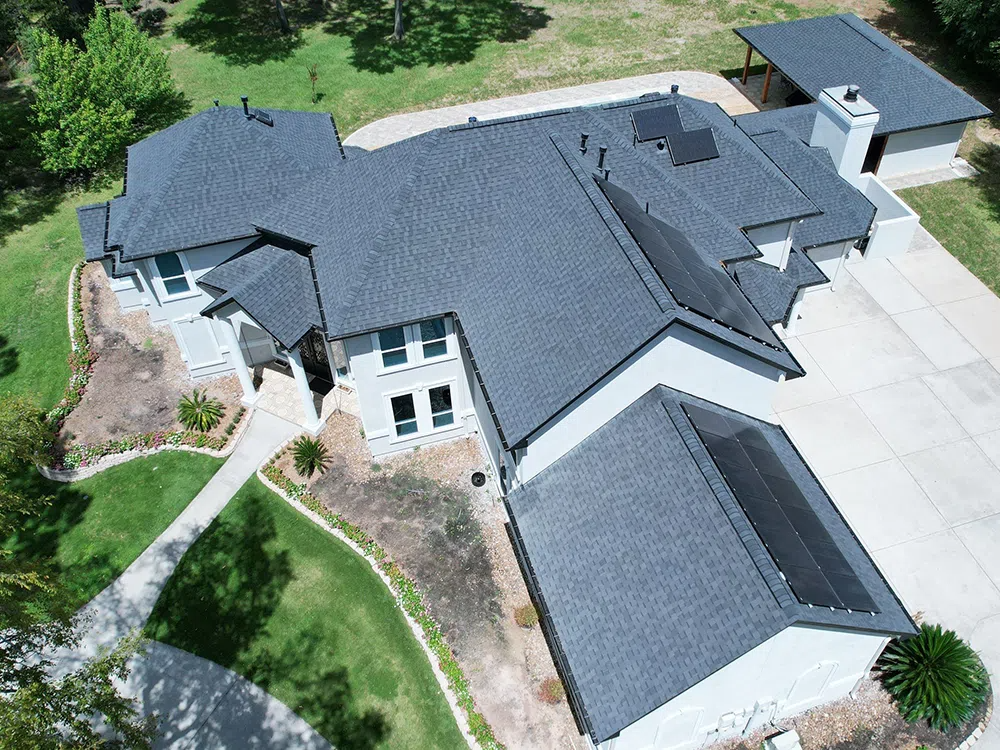 Aerial view of a gray-roofed, modern house with solar panels, a driveway, and surrounding greenery.