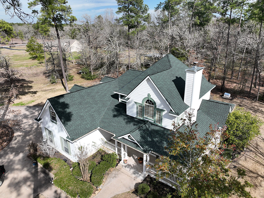Aerial view of a white house with a green roof, surrounded by trees and a long driveway.