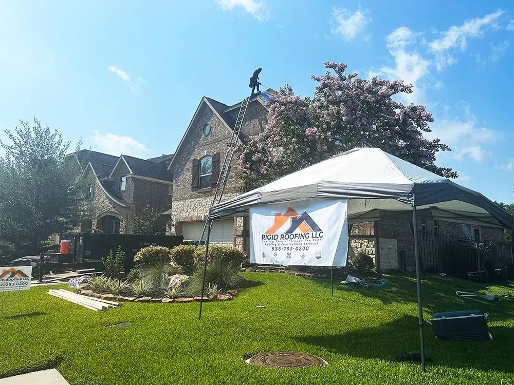 A person on a roof works on a house. A canopy and sign are in the yard, likely related to roofing services.