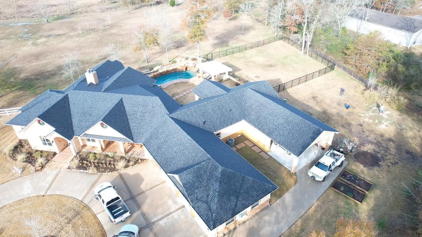 Aerial view of a large house with a dark roof, pool, and multiple vehicles in the driveway.