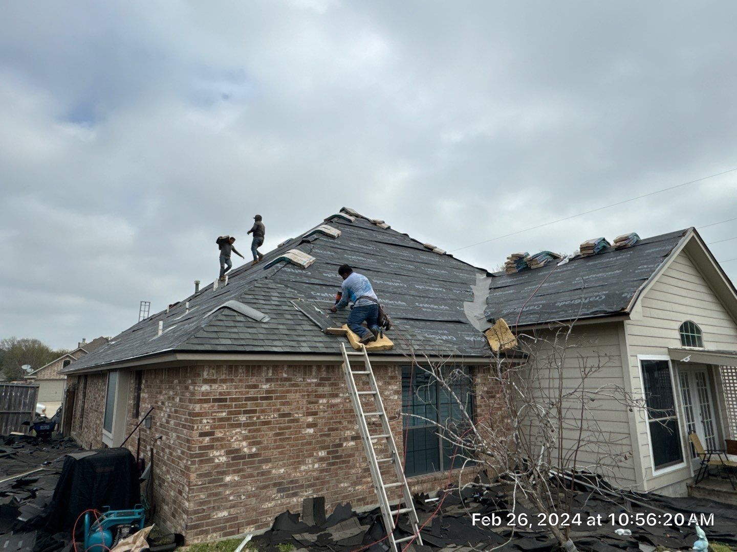 Three workers on a roof, removing old shingles. A ladder is propped against the roof. Cloudy sky overhead.