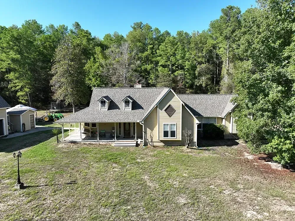 Tan house with porch and dormers sits in a grassy yard, surrounded by trees under a blue sky.