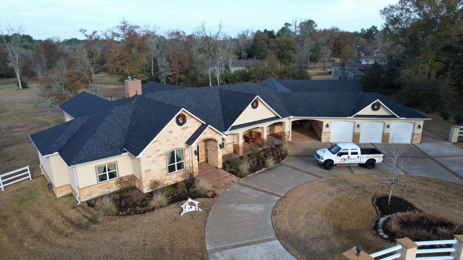 Aerial view of a large, tan-colored house with a dark roof and a circular driveway. A white truck is parked in front of the garage.