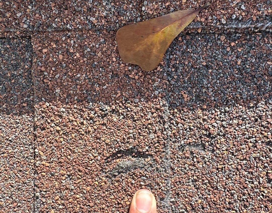 Close-up of brown roof shingles with a leaf and finger pointing at a damaged area.