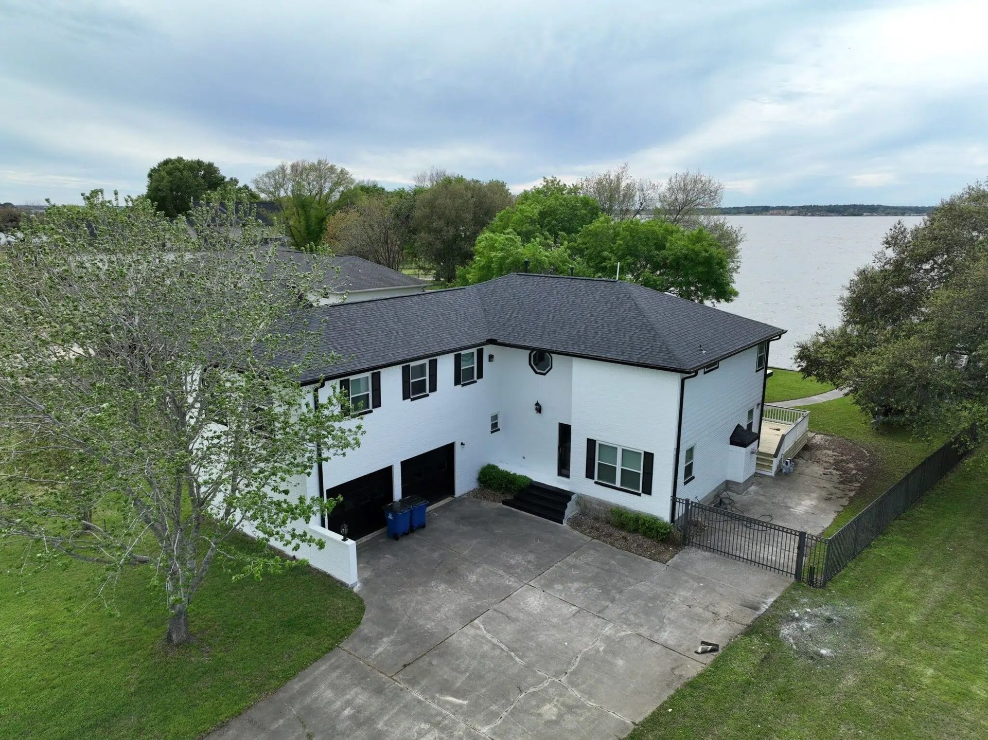 White two-story house with black shutters, garage, and roof, located by a lake under cloudy skies.