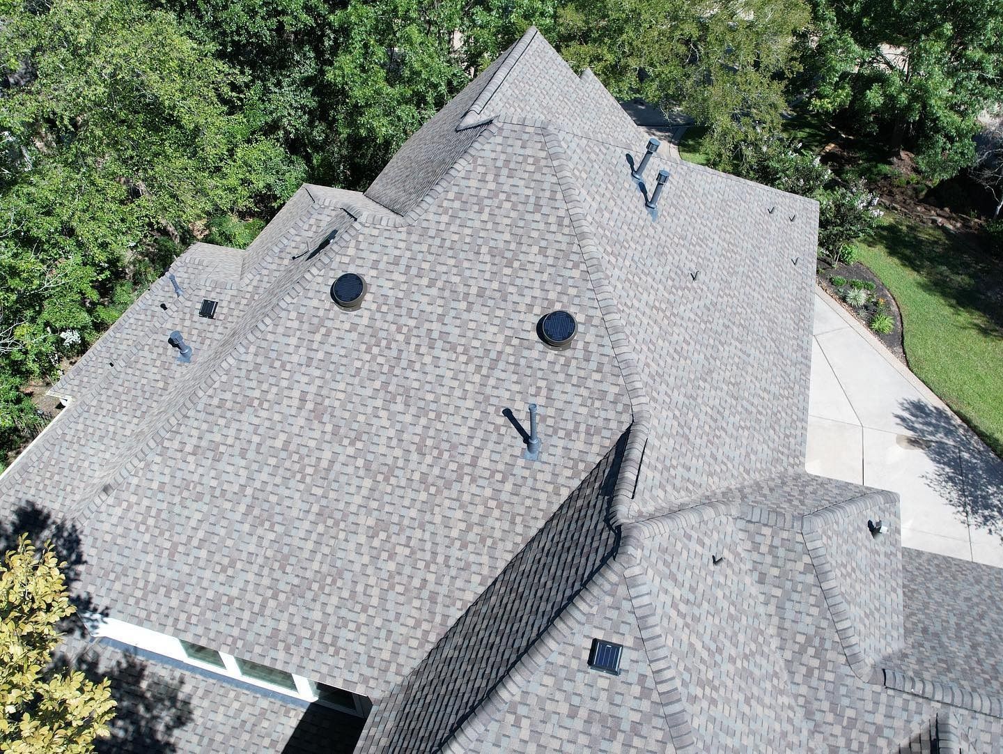 Overhead view of a house roof with asphalt shingles; surrounded by green trees.