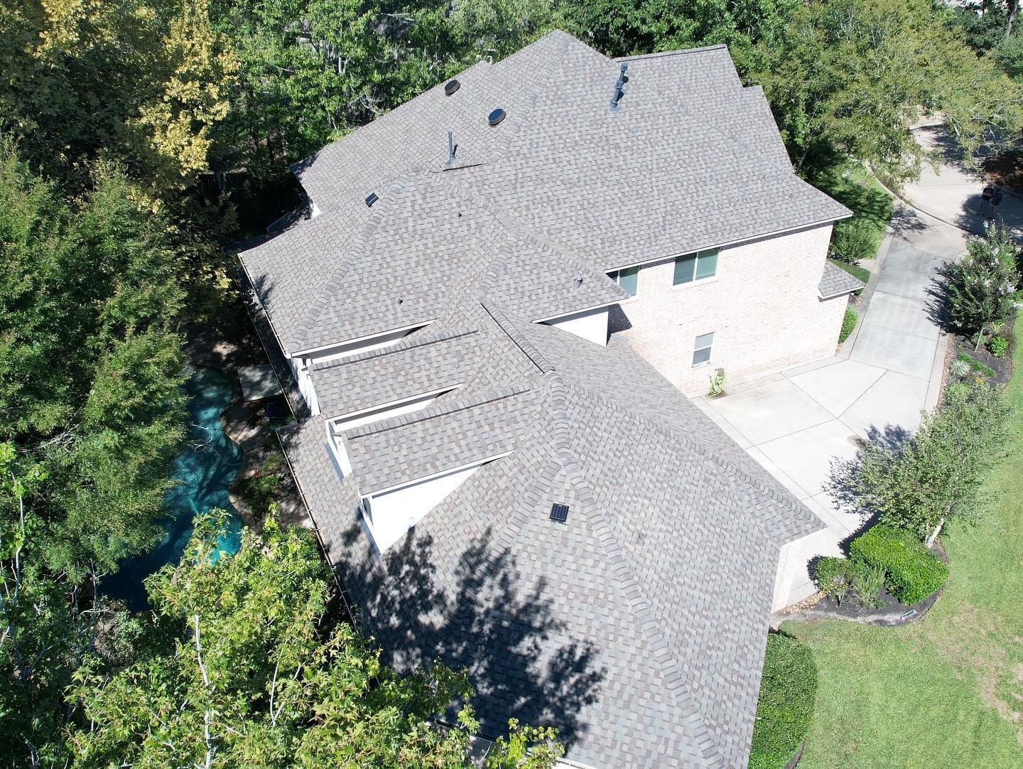 Aerial view of a two-story house with a gray shingled roof, surrounded by green trees and a driveway.
