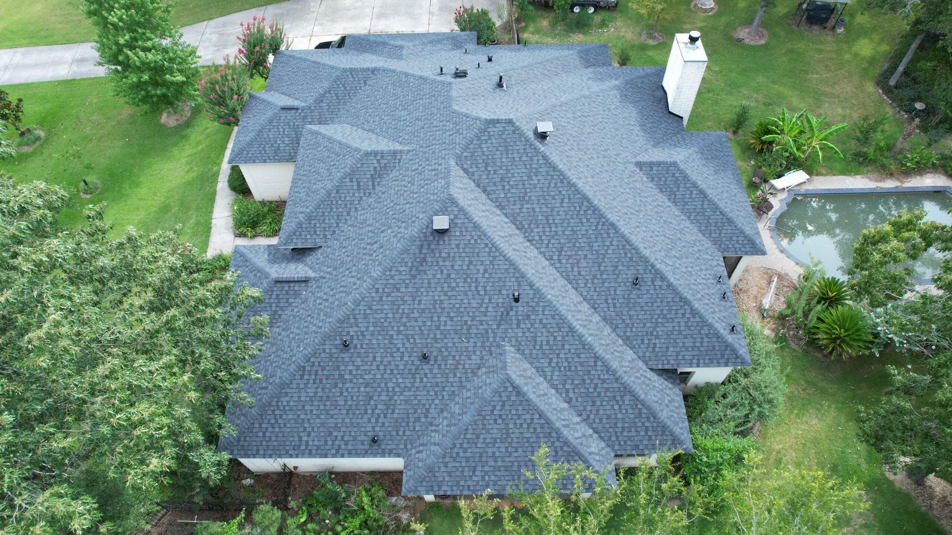Aerial view of a dark gray shingle roof of a house, surrounded by green trees, grass, and a pool.