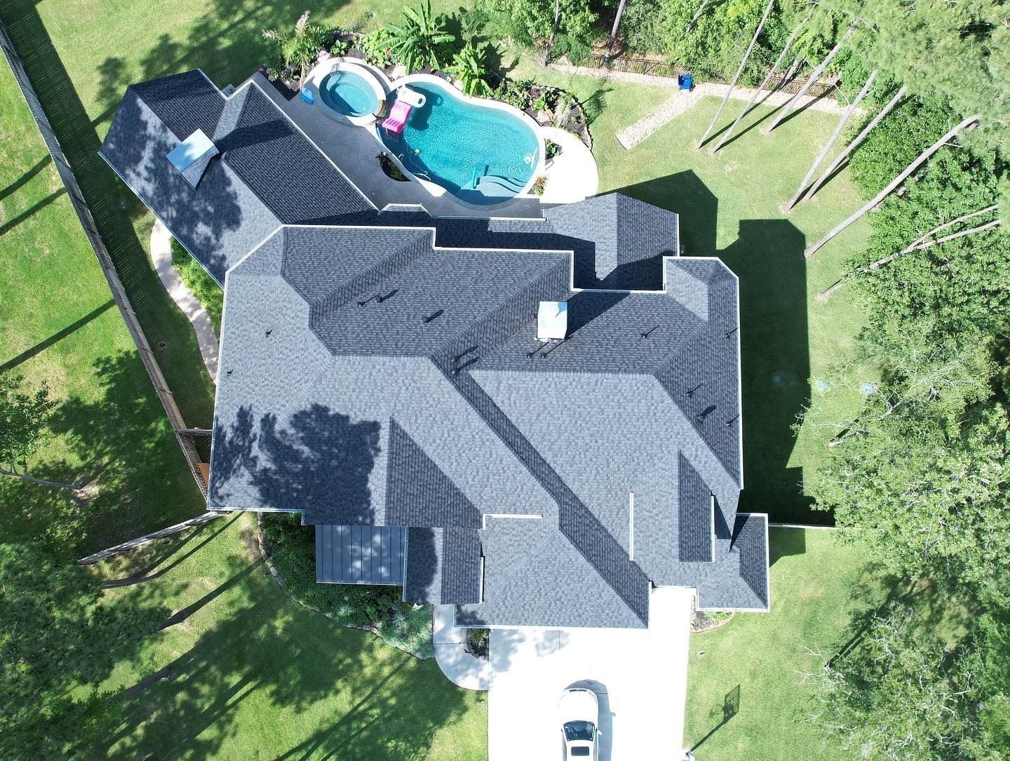 Aerial view of a dark-roofed house with a pool in a grassy yard, driveway and partial fence visible.