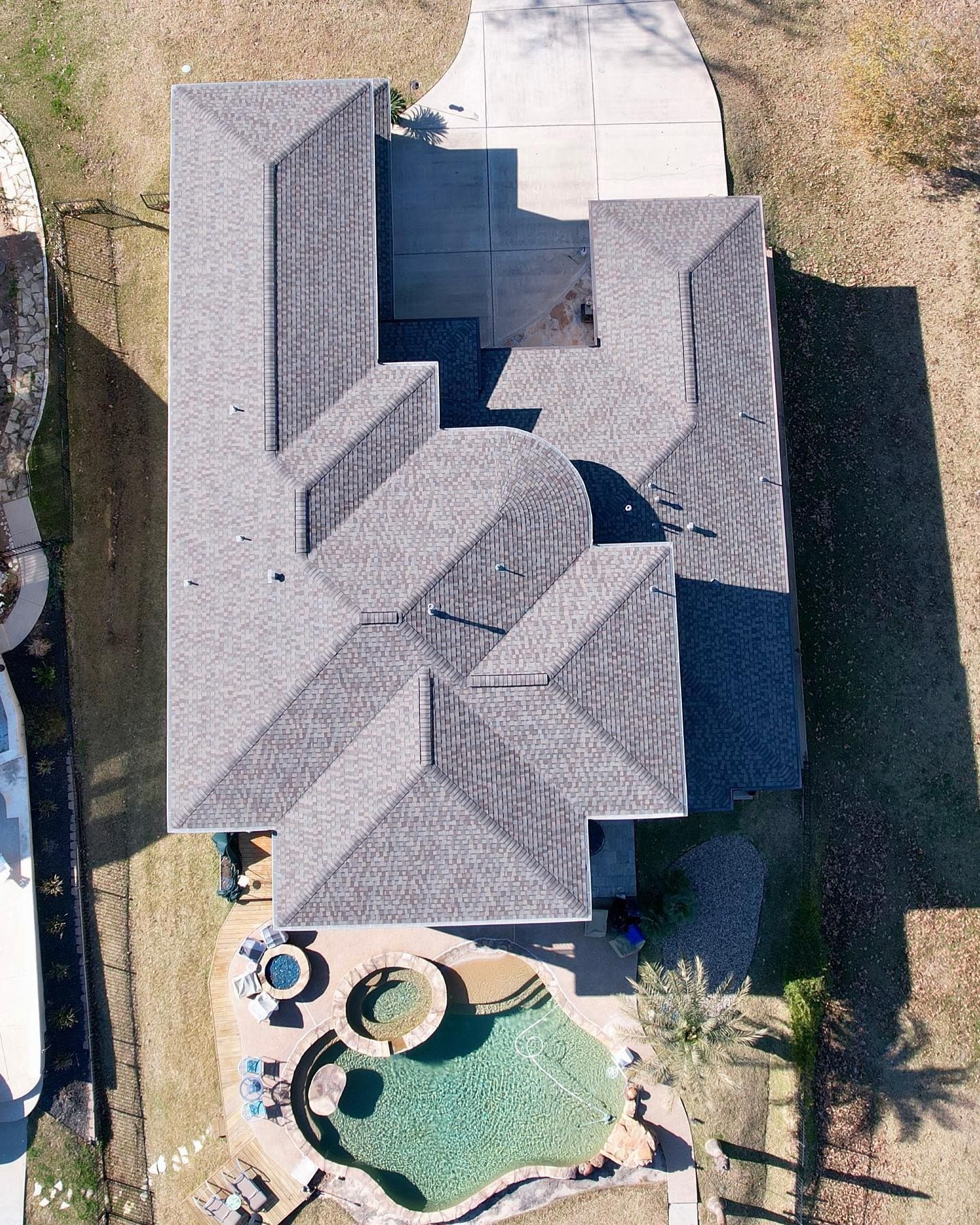 Aerial view of a house with a complex gray roof, driveway, and a pool in the backyard.